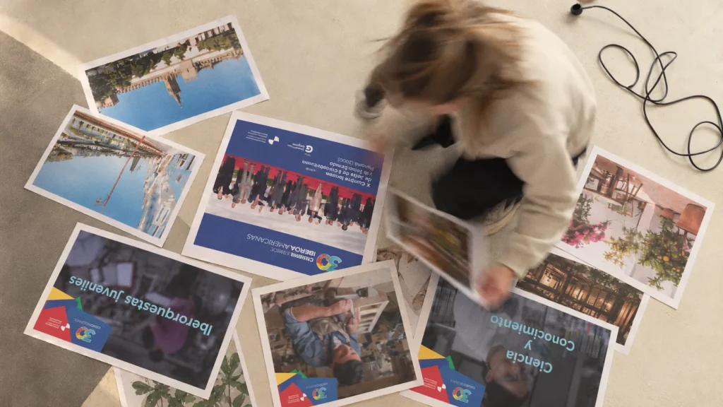 Vista cenital de una mujer, en movimiento, que revisa en el suelo las piezas impresas de la campaña de los 30 años de las Cumbres Iberoamericanas y fotografías de Sevilla